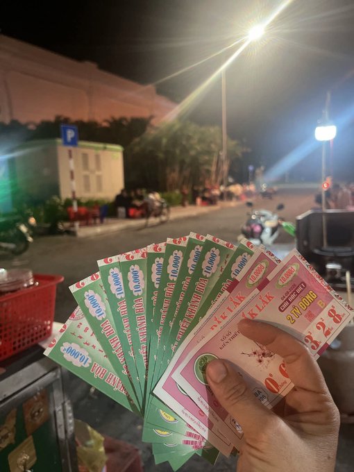 A hand holding a fan of Vietnamese lottery tickets at night on a street, with bright streetlights and blurred buildings and motorbikes in the background.