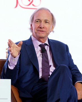 A man with light hair wearing a blue suit, lavender shirt, and dark tie, gesturing while speaking during what appears to be an interview.