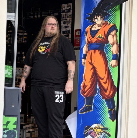 A man with long hair, glasses, and tattoos poses beside a large Dragon Ball Goku poster, wearing a black t-shirt and pants marked with "JUST DO IT 23." The image ties to the $PIERRE meme narrative by visually embodying the "French Goku Hero" character — the muscular anime figure beside him symbolizing the meme's inspiration drawn from French internet culture and viral Goku imagery.