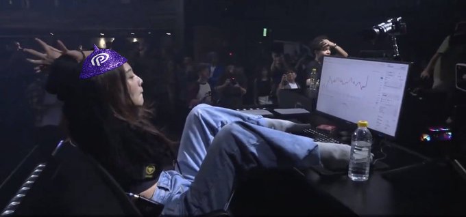 A streamer wearing a purple headband with a 'P' logo lounges with their feet up on a desk, looking at a monitor displaying a downward-trending line chart during what appears to be a live event.