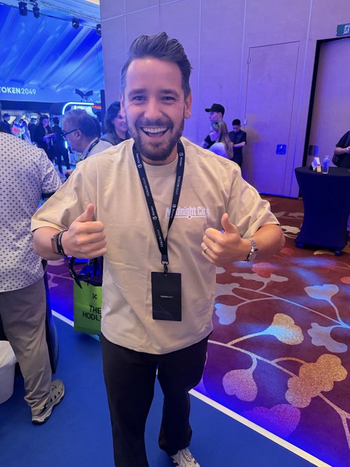 A smiling bearded man in a beige 'Foresight One' t-shirt giving a double thumbs-up at what appears to be a TOKEN2049 conference, wearing a lanyard with a black badge and a green 'THE HOOD' tote bag visible nearby.