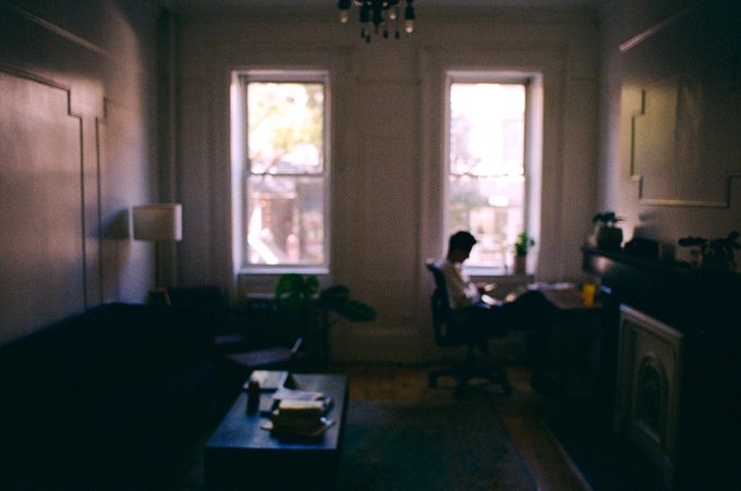 A dimly lit, atmospheric living room with two large windows, where a person sits at a desk working, illuminated by the natural light from outside.
