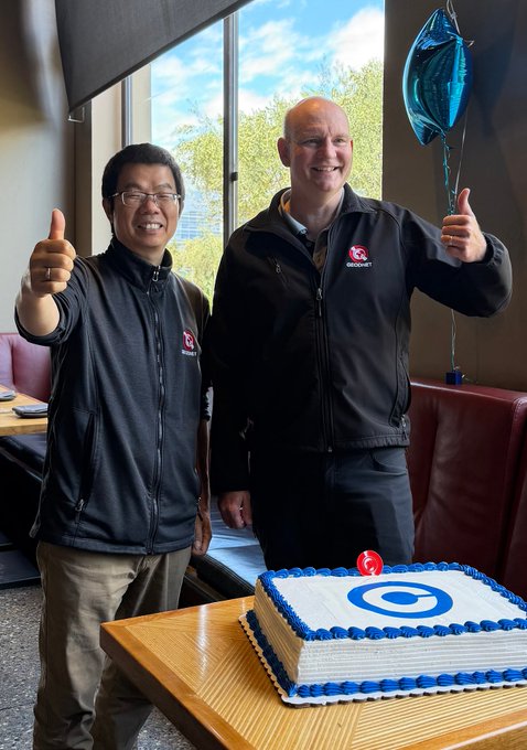 Two smiling men in GEODNET-branded jackets give thumbs-up behind a celebratory cake decorated with the GEODNET logo, marking the milestone of GEOD being listed on Coinbase. A blue balloon adds to the festive atmosphere as the GEODNET team pauses to recognize the community effort behind the achievement.