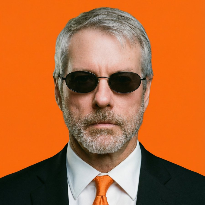 A portrait of an older man with gray hair and a beard, wearing dark oval sunglasses, a black suit, white shirt, and orange tie against a solid orange background.