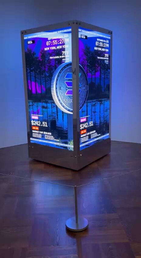 The image features a large, illuminated display screen that shows an advertisement for Bitcoin. The screen displays a coin with a blue background and white text, which reads "I miss the bull market vibes Will $sol ever get smash back to $240+?". The advertisement also includes a price of $115k+ for the coin. The display is situated on a wooden floor in front of a wall, creating an interesting visual contrast between the natural material and the digital technology.