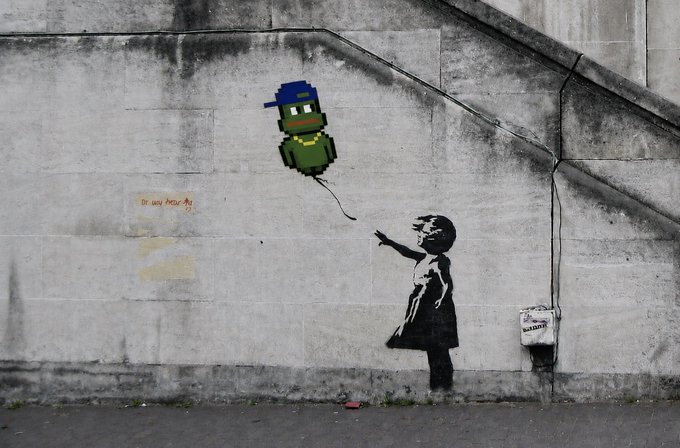 A photograph of street art on a weathered concrete wall, depicting a black-and-white stencil of a girl reaching toward a pixel-art green alien/zombie with a blue cap.