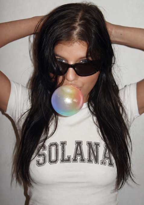 A woman with long dark hair wearing sunglasses and a white t-shirt reading "SOLANA" blows a colorful bubblegum bubble.