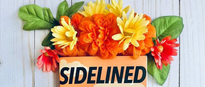 A floral arrangement of orange, yellow, and red flowers with green leaves sits behind an orange card displaying the word 'SIDELINED' in bold black text on a wooden surface.
