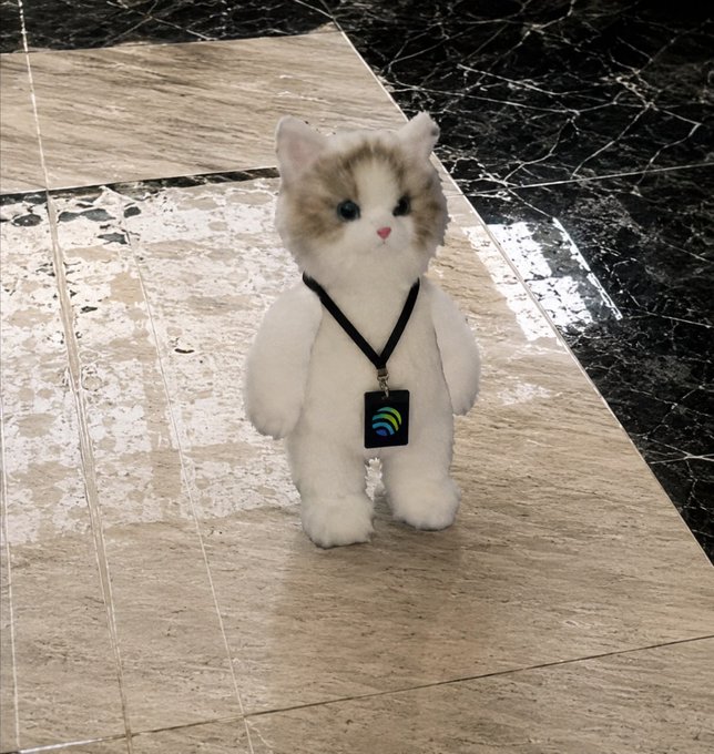 A small plush kitten toy with cream and white fur standing upright on a marble floor, wearing a black lanyard with an ID badge.