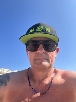 A shirtless selfie of a man wearing sunglasses and a black and neon green 'HATFISHING' cap with a blue sky background.