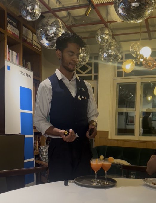 A bartender in a vest prepares two pinkish cocktails on a tray inside a stylish venue with disco balls and warm lighting, while a laptop displays "Stay Based" branding—tying into the post's appreciation for @base and the "stay based" theme.