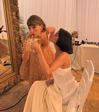A woman in a strapless white gown sits before an ornate gold mirror while another woman in a textured orange dress holds up a drink and appears to be adjusting her friend's hair, suggesting preparations for a formal event like a wedding. The image playfully visualizes the Polymarket speculation about Selena Gomez potentially serving as a bridesmaid at Taylor Swift's wedding, with the elegant styling evoking bridal party preparations.