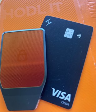 A square orange and blue device with a lock icon sits next to a blue Visa Debit card on an orange surface, appearing to be unboxing or product photography for the HODL11 hardware wallet.
