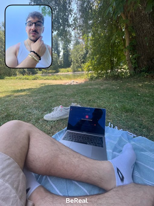 The image features a man sitting outdoors with his legs crossed, wearing white socks and shorts while using an open laptop computer placed on a blanket or towel next to him. The background includes trees and grass, creating a serene atmosphere for working or studying outside. The caption "bereal" is also present in the image.
