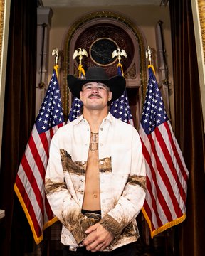 The image features a man wearing a cowboy hat, shirtless, and holding his hands together with his elbows resting on his knees. He stands next to two American flags that are placed behind him. The caption of the image reads "Our first athletes have already arrived at the White House! #UFCWhiteHouse coverage starts at 9 PM on @ParamountPlusBR [ UFC Freedom 250 presented by @cryptocom and @RamTrucks]". This suggests that the man is attending a sports event or gathering related to American football, possibly at the White House.