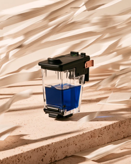 A product photograph of a transparent black ink cartridge or printer component filled with blue liquid, set against flowing beige fabric with dramatic lighting.