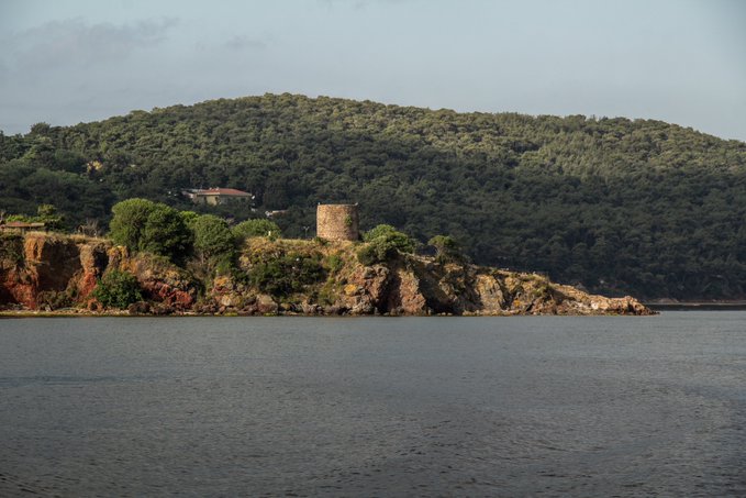 An old stone tower sits atop a rocky cliff surrounded by dense forest, viewed from across a calm body of water.