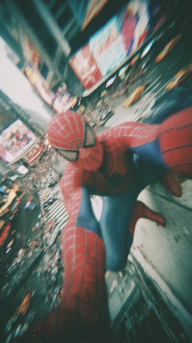 Spider-Man crouches in his iconic red and blue suit atop a building overlooking a bustling city street, with blurred billboards and yellow taxis visible below.