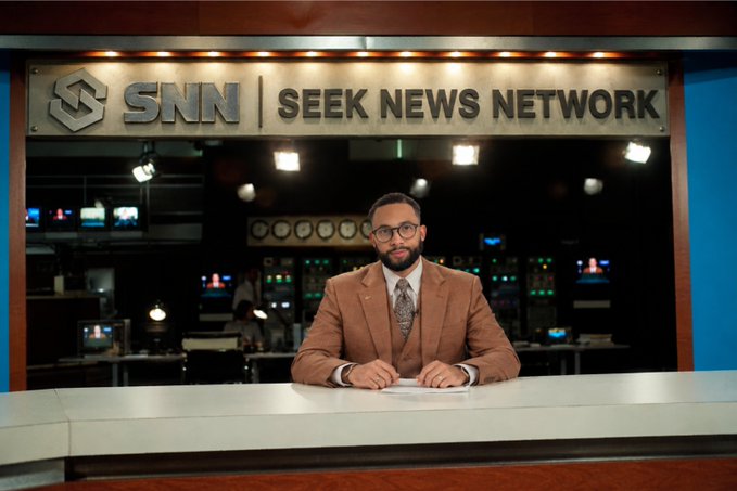 A male news anchor wearing glasses, a brown suit, and patterned tie sits at a desk in the SNN (Seek News Network) studio.
