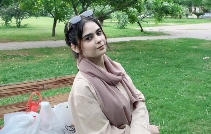 A young woman wearing sunglasses on her head and a beige hijab sits on a wooden park bench with shopping bags beside her, enjoying a sunny day in a lush green park. The image ties to the post as a relatable moment of post-trading leisure, celebrating weekend shopping sprees funded by weekly profits.
