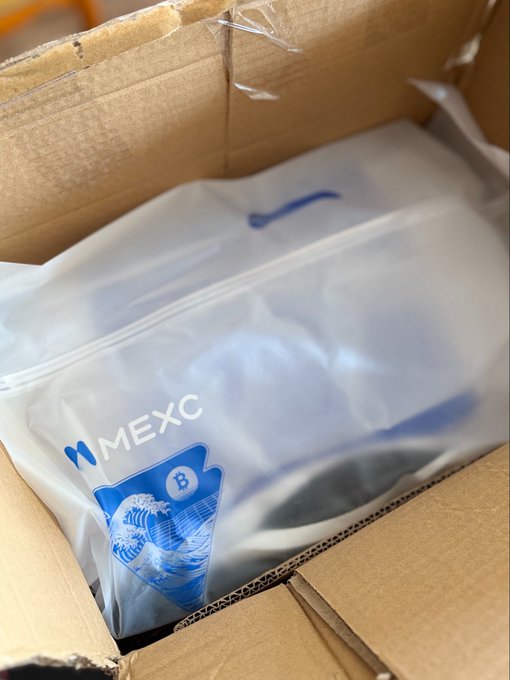 A cardboard shipping box contains a white plastic MEXC-branded bag with a Bitcoin logo holding some items.