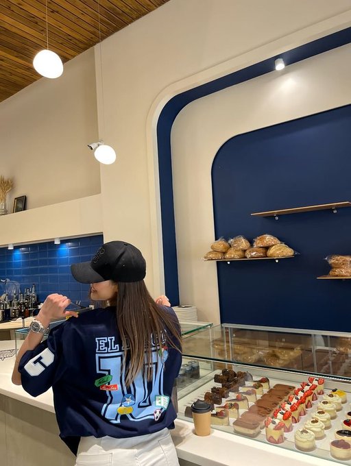 A person wearing a navy blue jersey with 'EL 51' on the back and a black cap stands at a pastry counter, looking at shelves of breads and a glass display case filled with various desserts.