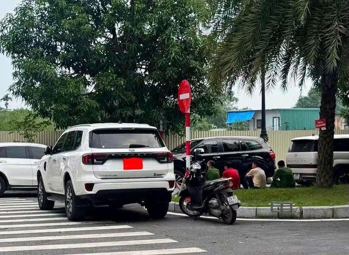 A street scene showing a white Ford SUV and other vehicles parked near a no-entry sign, with several people sitting on the grass beside the road.