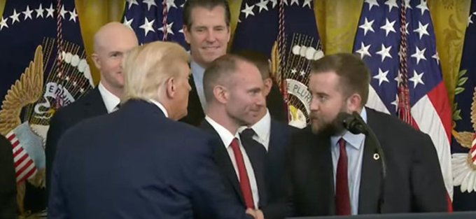 President Donald Trump with blonde hair is seen in profile at what appears to be a White House event, surrounded by several men in suits, with American flags displayed in the background.