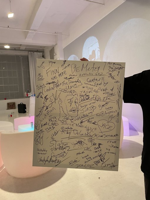 A large canvas or sign densely covered with various signatures and handwritten messages is held up in a modern, minimalist indoor space with white seating.