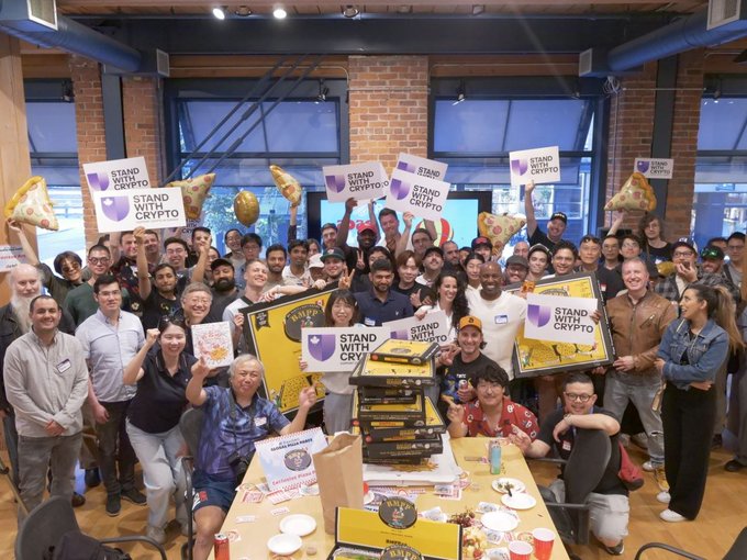 The image shows a large group of people gathered around a table filled with pizza boxes and signs promoting Bitcoin. The atmosphere appears festive as everyone seems to be enjoying themselves. The event has been organized by @standwithcrypto, an organization dedicated to raising awareness about cryptocurrency issues. The attendees are holding up signs that read "Stand with Cryptocurrency" and "PizzaDAO celebrated our 6th Bitcoin Pizza Day on Friday". This gathering is a celebration of the progress made in the world of cryptocurrencies and the support for this emerging technology among the community.