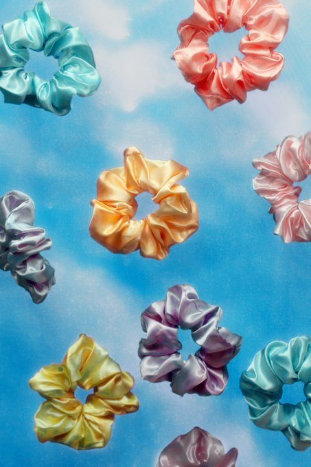 An artistic composition of pastel-colored satin scrunchies in shades of teal, pink, orange, lavender, and yellow scattered against a soft blue cloudy background.