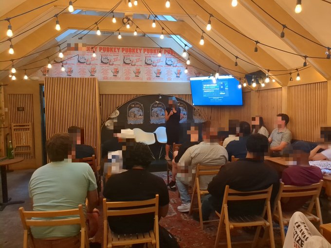 The image shows a group of people sitting in chairs around a table, with some standing as well. A woman is giving a presentation or lecture while others listen attentively. The room has wooden walls and ceiling, creating an intimate atmosphere for the gathering. In the background, there are signs that read "Pubkey Puffin" and "@Pubkey NYC Privacy Pop-up", indicating that this event might be related to privacy concerns in the digital age or a discussion about blockchain technology.
