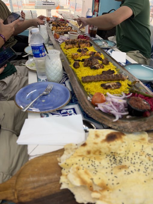 A large wooden platter of yellow rice with meat, served family-style at a restaurant, sits on a table with fresh flatbread, onions, tomatoes, water bottles, and blue plates, while diners reach in to serve themselves.