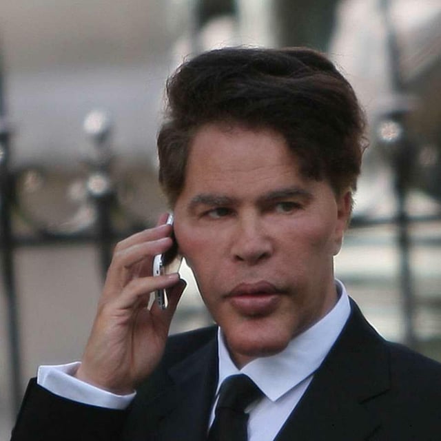 A man with dark hair wearing a black suit, white shirt, and black tie, holding a flip phone to his ear on a city street.
