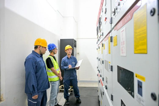 The image shows a group of three people standing next to each other inside an industrial setting with white walls and yellow signs. One person appears to be wearing a hard hat, indicating that they are likely construction workers or engineers. The man holding papers is talking to his colleagues about their work on the project. The post in front of them reads "Shantou, Guangdong taps new growth momentum via AI token exports, delivering high-value AI computing services to overseas users through its international submarine cables." This suggests that they are discussing a project related to artificial intelligence or technology and how it can be applied to their work.