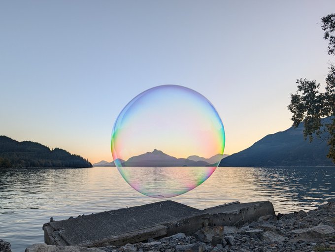 A large, iridescent soap bubble floats in front of a scenic mountain lake at sunset, with a piece of concrete debris in the foreground.
