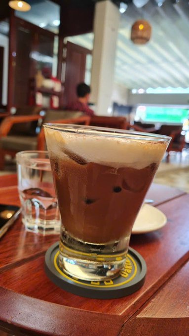 A tall glass of iced coffee with a thick foam layer sits on a coaster on a wooden café table, with a glass of water beside it and a blurred interior in the background.