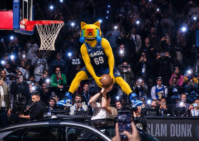 A person in a yellow cat/dog mascot costume wearing sunglasses and a Magic #69 jersey dunks a basketball over a car at an NBA slam dunk contest, surrounded by a crowd with flash photography.