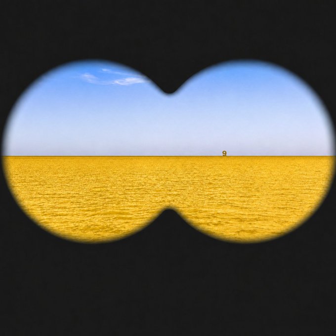 The image shows a view through binoculars of a yellow field beneath a blue sky, with the number "9" visible on the horizon.

The post mentions a "GM Eye Test Challenge" asking viewers to find a hidden character. The two circular binocular lenses cleverly form the outline of a face, and the small "9" on the horizon represents the nose, while the curved bottoms of the lenses create a smiling mouth — revealing a hidden happy face when viewed through the binoculars' perspective.