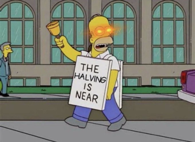 A Simpsons-style character stands on a sidewalk holding a bell and a sandwich-board sign reading "THE HALVING IS NEAR," serving as a humorous prophetic messenger tied to the post's announcement that the $BTC halving is coming.