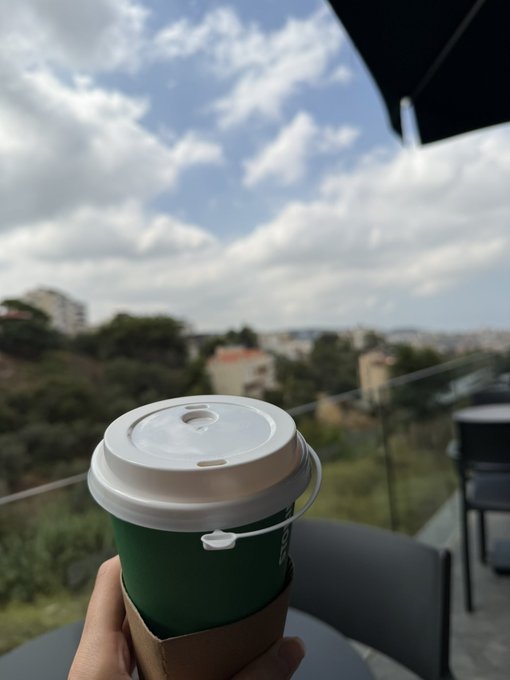 A hand holds a green paper coffee cup with a white lid against a backdrop of a cloudy sky, distant city buildings, and trees.