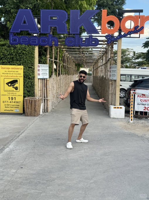 A man in a black sleeveless shirt, beige shorts, and sunglasses posing with open arms in front of the entrance gate to ARK bar beach club, with CCTV warning signs and a Pakin customs sign visible.