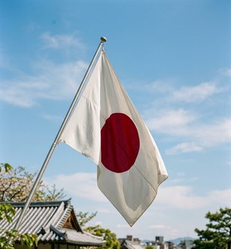 A photograph of the Japanese national flag (white with a red circle) waving on a flagpole against a bright blue sky, with traditional rooftops and green trees visible in the background.
