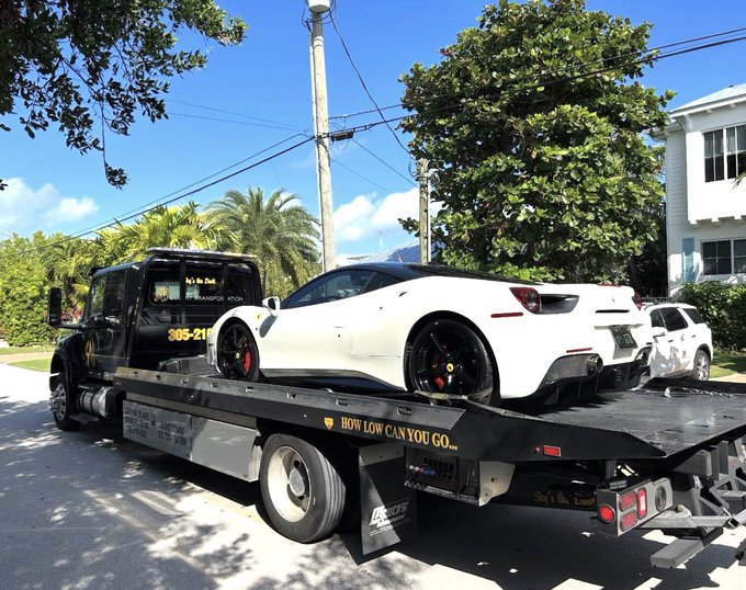 A white sports car with red brake calipers being transported on a flatbed tow truck, which features a humorous bumper sticker reading 'HOW LOW CAN YOU GO...'.