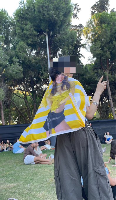 A person wearing a green and yellow striped scarf and cargo pants poses with a peace sign at an outdoor event, fitting the post's "if you see green, that's us tm" theme of group identification through color. Other attendees sit on the grass in the background, with trees surrounding the venue.