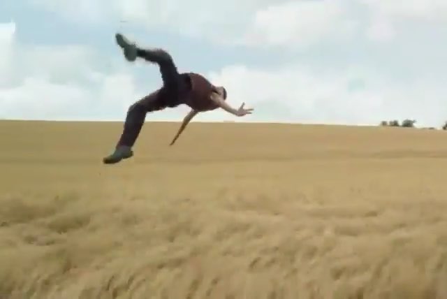 A person is captured mid-air, seemingly tumbling or falling backward over a golden wheat field under a partly cloudy sky.