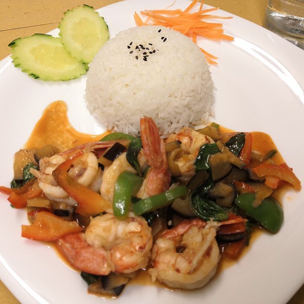 A white plate holds a serving of white rice topped with sesame seeds, a shrimp stir-fry with vegetables in a reddish sauce, cucumber slices, and shredded carrots.
