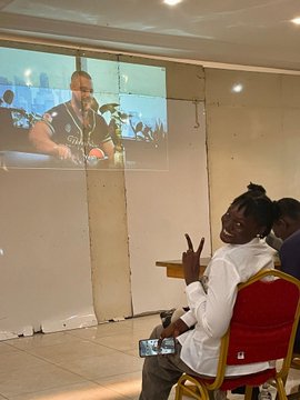 The image shows two people sitting next to each other, one person wearing a white shirt and the other wearing a red chair. They are both smiling while watching something on a projection screen or wall hanging behind them. The room they're in has a tiled floor and a white wall with a projector screen mounted on it. There is also a cell phone visible near the bottom of the image, possibly used to capture memories from their time together.