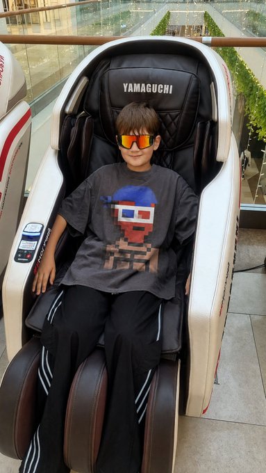 A child wearing reflective orange sunglasses and a gray graphic t-shirt sitting in a "YAMAGUCHI" branded massage chair in what appears to be a mall.