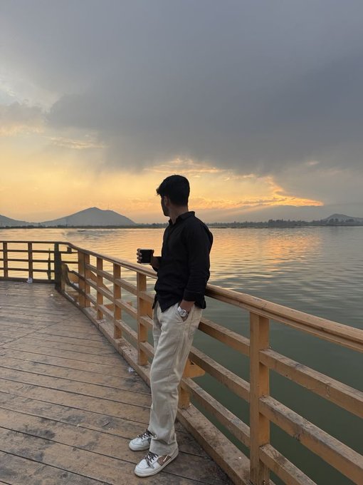 A man in a dark shirt and light pants stands on a wooden pier holding a cup of coffee, looking out at a calm body of water under a dramatic sunset sky.
